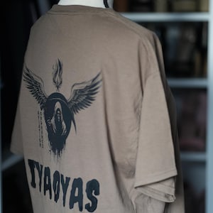 Op de afbeelding: Een beige t-shirt met een zwarte afbeelding op de achterkant. Het ontwerp toont een gevleugelde figuur met een zeis en de tekst "TYAOAYAS". Het ontwerp bevat de woorden "ORDNANCE AVIATION".