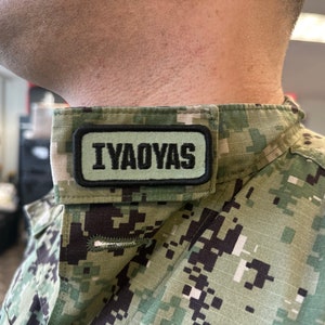 Puede incluir: Una camisa de uniforme militar con un patrón de camuflaje. Un parche rectangular con el texto "IYAOYAS" en letras negras está cosido en el cuello.