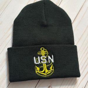 以下が含まれることがあります： 黒色のニット帽。折り返されたつばがあり、前面には黄色と白の刺繍された錨のデザインと、錨の上に「USN」の文字があります。 この帽子は柔らかいニット素材で作られています。