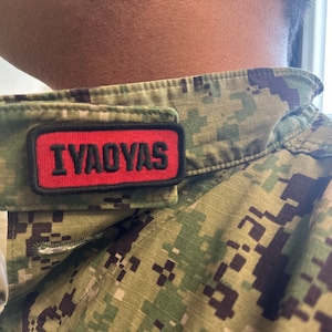 May include: Close-up of a military uniform with a camouflage pattern. A red patch with the word "IYAOYAS" in black letters is sewn onto the uniform's collar. The uniform is a mix of green, brown, and black.