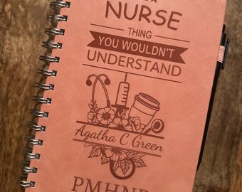 Op de afbeelding: Een zalmkleurig spiraalgebonden notitieboek met de tekst "IT'S A NURSE THING YOU WOULDN'T UNDERSTAND." Het notitieboek bevat een stethoscoop, spuit, koffiekopje en bloemenontwerp. De naam "Agatha C Green" en "PMHNP" staan ook op de kaft.