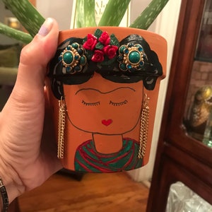 May include: A terracotta flower pot painted to look like Frida Kahlo. The pot has a black painted hairstyle, red roses, and turquoise blue earrings. The pot is painted with a brown face, closed eyes, and a red heart-shaped mouth.