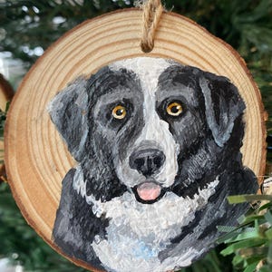 Op de afbeelding: Ronde houten kerstversiering met een handgeschilderd hondenportret. De hond heeft zwart-witte vacht, gele ogen en zijn tong is zichtbaar. Er is een touwlus bevestigd om op te hangen. De versiering hangt in een kerstboom.