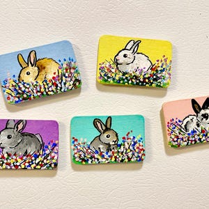 Puede incluir: Cinco rectángulos de madera pintados a mano, cada uno con una ilustración de conejo. Los conejos están representados en varios colores, sobre un fondo de flores coloridas y un cielo azul. Los rectángulos están pintados en tonos de azul, amarillo, morado, rosa y turquesa.