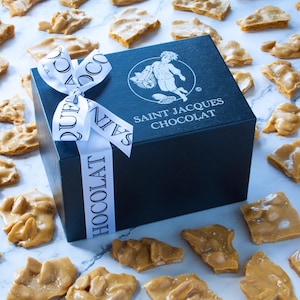 May include: A black gift box with a white ribbon and the text "SAINT JACQUES CHOCOLAT" is surrounded by pieces of peanut brittle. The box has a logo of a cherub carrying a basket. The ribbon has the word "CHOCOLAT" printed on it.
