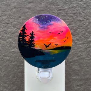 Puede incluir: Luz de noche redonda con un diseño de atardecer. Presenta árboles silueteados, pájaros volando y un lago. El cielo es una mezcla de morado, rosa y naranja. La luz de noche tiene una base blanca.