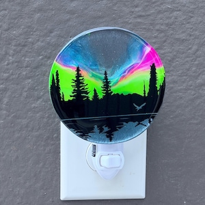 May include: A circular night light featuring a vibrant resin art design of a neon green and pink aurora borealis above a black silhouette of a pine forest and a lake. A small bird is visible in flight, and the light is plugged into a white wall outlet.