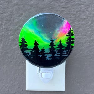 May include: A circular night light featuring a vibrant resin design of a neon green and pink aurora borealis sky above a silhouette of black pine trees. The light is mounted on a standard white plastic plug-in base with a manual toggle switch.