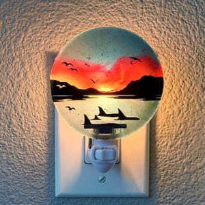 Op de afbeelding: Een rond glazen nachtlampje met een zonsonderganglandschap, een rode en oranje lucht, donkere bergsilhouetten en zwarte contouren van vogels en dolfijnen in het water, aangesloten op een standaard wit stopcontact in de muur.