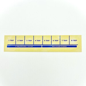 May include: A yellow measuring guide with blue markings for tablespoons. The guide shows 1 to 8 tablespoons, with the text "4 tablespoons = 3/4 cup" and "Wallflour Studios" printed on it.