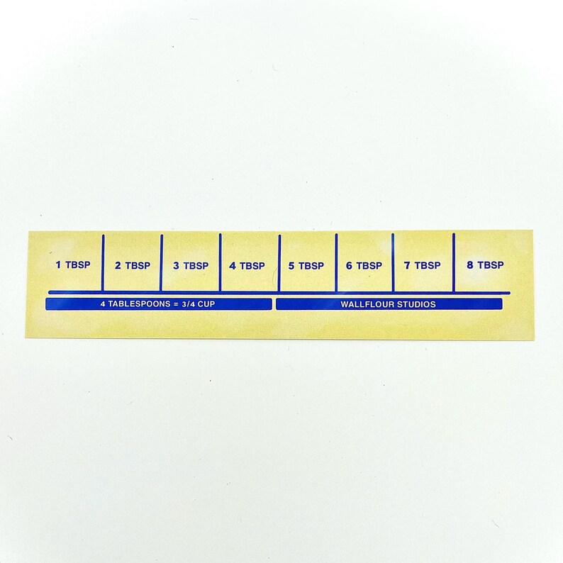 May include: A yellow measuring guide with blue markings for tablespoons. The guide shows 1 to 8 tablespoons, with the text "4 tablespoons = 3/4 cup" and "Wallflour Studios" printed on it.