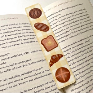 May include: A bookmark with a yellow and white checkered background and various brown bread illustrations. The bookmark features text from a book about a woman who befriended ghosts. The text reads "The woman who befriended ghosts... seemed she had failed. You can't force people to be friends..."