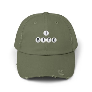 May include: Olive green distressed baseball cap with the text "I BITE" spelled out in white letter tiles. The cap has a curved brim and a distressed, worn look.