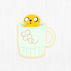 May include: A yellow cartoon dog with brown eyes is peeking out of a light green mug with a pink flower design. The mug has a handle and a light brown rim.