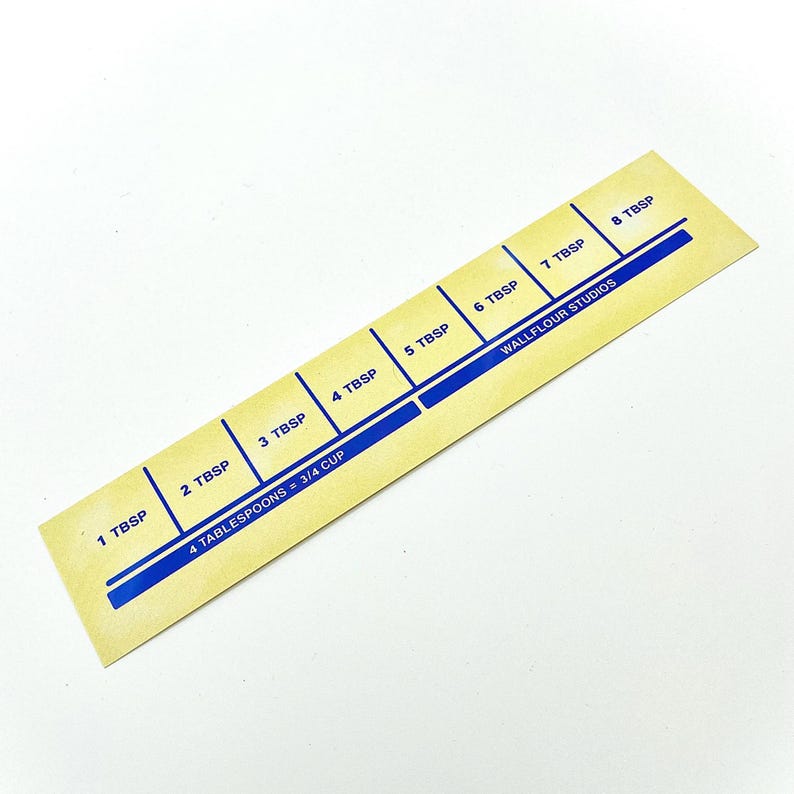 May include: A yellow measuring tool with blue markings for tablespoons. The tool is labeled "4 tablespoons = 3/4 cup" and "Wallflour Studios".