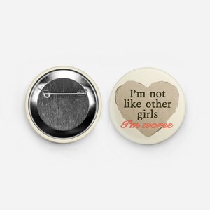 May include: Two round cream-coloured pinback buttons. One button shows the back with a safety pin. The other button has a heart-shaped cutout revealing a brown background. Text reads "I'm not like other girls I'm worse."