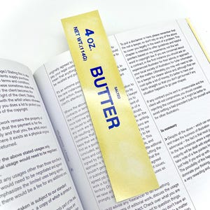 May include: A yellow bookmark with the words "SALTED BUTTER" in blue lettering and "4 oz. NET WT.(114G)" in black lettering. The bookmark is made of a glossy material and has a gold sheen.