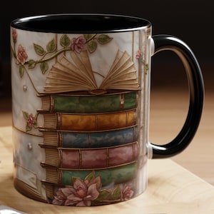 May include: A ceramic coffee mug with a black handle and interior. The mug features a stack of books with an open book on top, adorned with floral and vine details. The mug is on a wooden surface.
