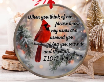 May include: A round, glass Christmas ornament with a red cardinal perched on a snow-covered branch. The ornament features a white background with a message that reads: "When you think of me, please know my arms are around you giving you warmth and strength. I LOVE YOU!"