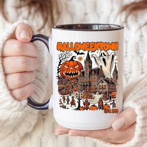 May include: White ceramic mug with a black rim and handle. The mug features a colorful illustration of a haunted house, a ghost, and pumpkins with the text "Halloweentown Est. 1999".