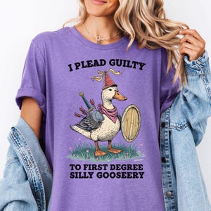 Op de afbeelding: Paars T-shirt met een cartoonillustratie van een eend die een kroon draagt en een zwaard vasthoudt. De tekst op het shirt luidt "Ik pleit schuldig aan eerstegraads domme ganzenstreken."