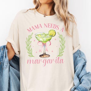 May include: A white t-shirt with the text "Mama Needs a Margarita" in pink lettering. A green margarita glass with a lime wedge is illustrated above the text.