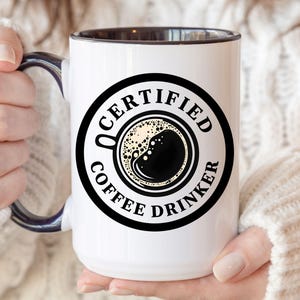 May include: White ceramic mug with a black rim and handle. The mug has a black and white graphic of a coffee cup with the text "CERTIFIED COFFEE DRINKER".