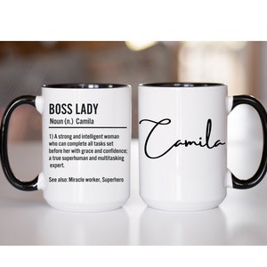 Może przedstawiać: Biały kubek ceramiczny z czarnym rantem i uchwytem. Kubek ma czarny wzór z napisem "Boss Lady" z definicją "Rzeczownik (n.) Camila" i listą atrybutów. Po drugiej stronie kubka znajduje się imię "Camila" napisane kursywą.