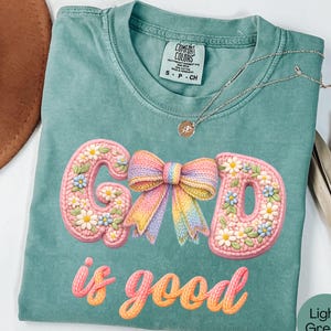 Puede incluir: Camiseta verde claro con la frase bordada "GOD is good". Las letras son rosas y están decoradas con flores blancas y azules. Un lazo colorido está entre las letras "G" y "D". Un collar plateado está sobre la camiseta.