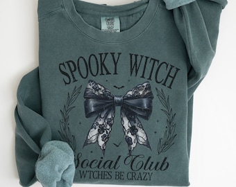 Könnte beinhalten: Ein tealgrüner Pullover mit dem Text "Spooky Witch Social Club" und einer schwarzen Schleife aus Spitze mit einem schwarzen Band. Der Text unter der Schleife lautet "Witches Be Crazy".