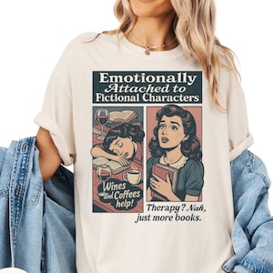 May include: Ivory-colored t-shirt with a retro comic-style graphic. The graphic reads "Emotionally Attached to Fictional Characters" and features illustrations of a woman with wine and books. The text "Wines and Coffees help!" and "Therapy? Nah, just more books." are also included.