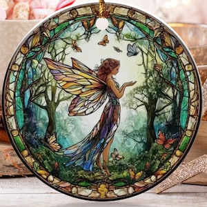 May include: Round ceramic Christmas ornament featuring a fairy in a forest scene with butterflies. The ornament has a gold string for hanging. Other holiday decorations include a gold star, a Christmas tree, and a decorative ball.