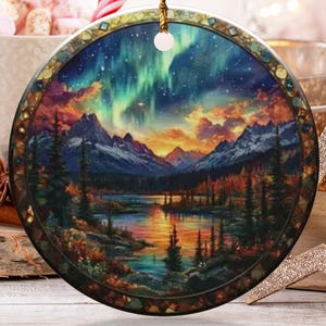 May include: Round ceramic Christmas ornament depicting a landscape with mountains, a lake, and the aurora borealis. The ornament has a gold rim and a gold hanging cord. Other decorations include a gold star, a gold glittery ball, and candy canes.