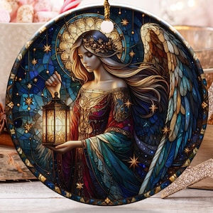 May include: A round Christmas ornament featuring a stained-glass style angel holding a lantern. The angel has long blonde hair and is wearing a colorful gown. The ornament is hanging by a gold string. Other holiday decorations are in the background.