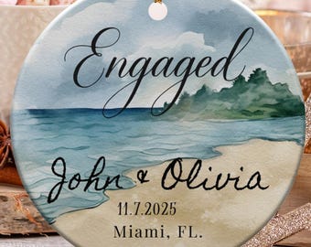 May include: Round ceramic ornament with a beach scene, the word "Engaged", and the names "John & Olivia" with a date and location. The ornament has a gold string for hanging. Other decorations include candy canes and a star.