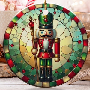 May include: A round Christmas ornament featuring a stained-glass design of a nutcracker soldier in red, green, and gold. The ornament hangs from a gold string. Other holiday decorations are in the background.