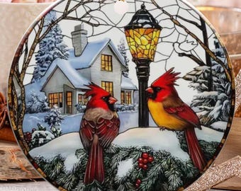 Peut inclure: Ornement de Noël rond avec une scène hivernale. Deux cardinaux rouges sont perchés sur une branche enneigée. L'ornement a une ficelle dorée. Autres décorations de Noël en arrière-plan.