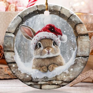 May include: Round Christmas ornament featuring a bunny wearing a Santa hat, set against a snowy backdrop within a stone archway. The ornament is suspended by a gold string. Other holiday decorations include candy canes and a star.