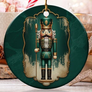 May include: A round Christmas ornament featuring a green and gold nutcracker design. The ornament is hanging by a gold string. The background includes a cup of marshmallows, candy canes, and other holiday decorations.