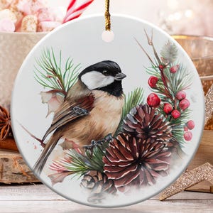 May include: A round white ceramic ornament hanging from a gold string, featuring a detailed watercolor illustration of a black-capped chickadee perched among green pine needles, brown pinecones, and clusters of small red berries.