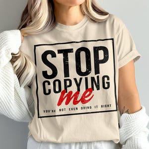 Puede incluir: Una camiseta beige con un gráfico cuadrado negro que dice "STOP COPYING ME YOU'RE NOT EVEN DOING IT RIGHT". El texto "ME" está en rojo.