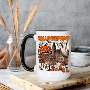May include: White ceramic mug with a black handle. The mug features a colorful illustration of a spooky town with a haunted house, pumpkins, ghosts, and the text "HALLOWEENTOWN EST. 1998".