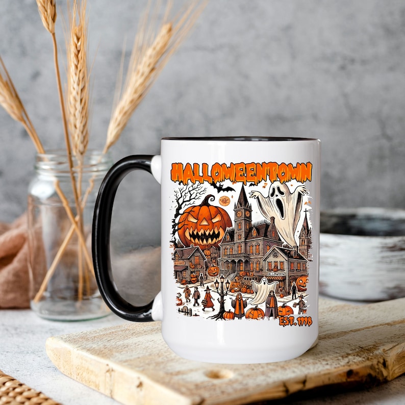 May include: White ceramic mug with a black handle. The mug features a colorful illustration of a spooky town with a haunted house, pumpkins, ghosts, and the text "HALLOWEENTOWN EST. 1998".