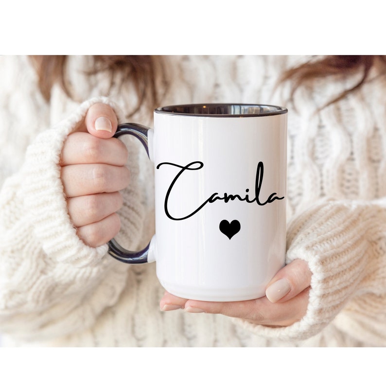 May include: A white ceramic mug with a dark blue interior and handle. The exterior features the name Camila written in a black cursive script font, positioned above a small, solid black heart icon centered on the front of the mug.