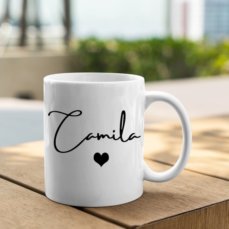 May include: A white ceramic mug featuring the name Camila printed in a black cursive script font. Below the name is a small, solid black heart icon. The mug has a standard cylindrical shape with a curved handle on the right side.