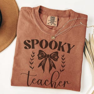 以下が含まれることがあります： テラコッタ色のTシャツ。ダークブラウンの筆記体で「Spooky teacher」と書かれています。「Spooky」の下には装飾的なリボンと葉の小枝がプリントされています。ペンダント付きのゴールドネックレスがシャツの上にかけられています。