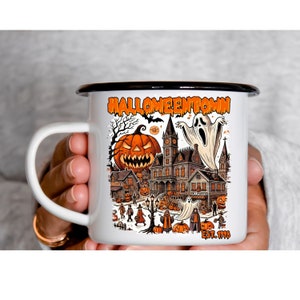 May include: White enamel mug with a black rim and handle. The mug features a colorful illustration of a spooky Halloween town with a haunted house, pumpkins, ghosts, and the text "Halloweentown Est. 1998".