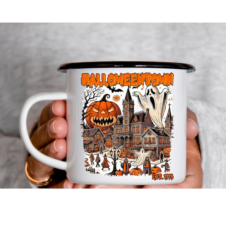 May include: White enamel mug with a black rim and handle. The mug features a colorful illustration of a spooky Halloween town with a haunted house, pumpkins, ghosts, and the text "Halloweentown Est. 1998".