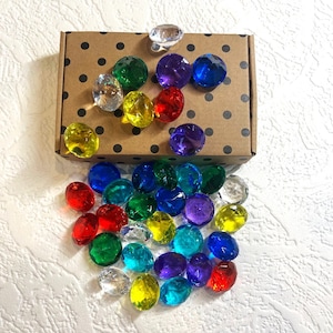 Puede incluir: Una caja marrón con lunares blancos llena de gemas de plástico de colores. Las gemas son de varios tonos de rojo, azul, verde, amarillo y transparente. Las gemas se derraman de la caja sobre una superficie blanca.