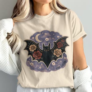 May include: Beige t-shirt featuring a black bat with floral wings, a crescent moon, and the text "Stay Weird." The bat design includes red and yellow flowers, set against a purple and blue cloudy background.